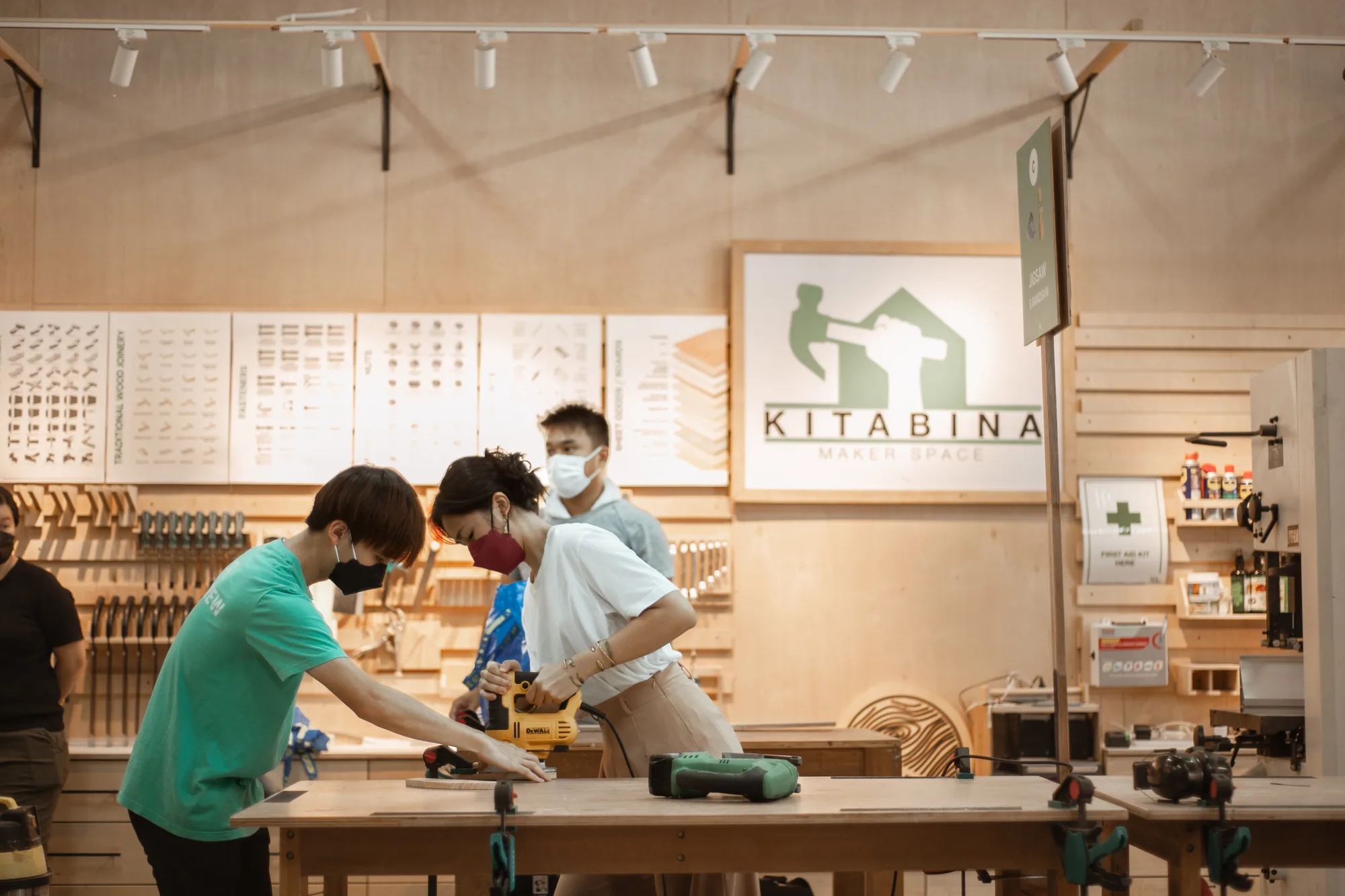 A KitaBina facilitator guiding a maker through a jigsaw cut at the bench, with others working alongside