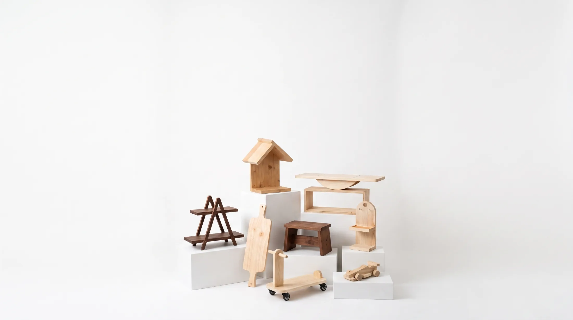 Nine finished Pesta Bina projects grouped on white plinths against a white studio backdrop: a birdhouse, an A-frame shelf, a cutting board, a stool, a balance board, a scooter, a toy car and a small arched shelf